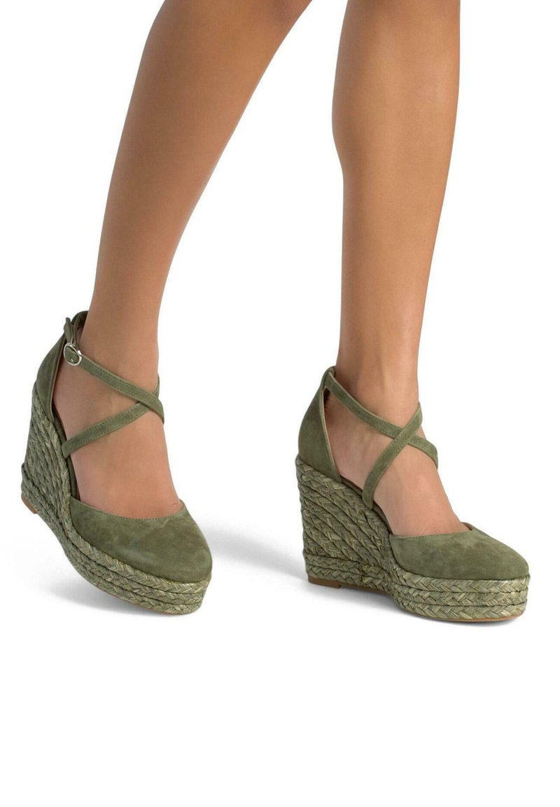 Zielone zamszowe koturny z krzyżującymi się paskami na kostce i teksturowanymi detalami z liny na szerokiej podeszwie. Gładka strefa palców, zaokrąglony kształt.