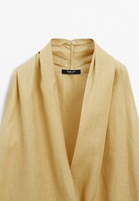 Haut en lin beige avec un décolleté en V profond, présentant un drapé doux et une étiquette discrète au dos. Tissu texturé avec une finition naturelle.