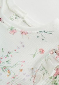 Tela blanca con un acabado texturizado que presenta un estampado floral en rosa, púrpura y verde. Incluye un detalle con volantes a lo largo del escote.