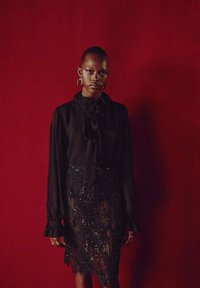 Blusa nera con fiocco e maniche lunghe, abbinata a una gonna di paillettes fino al ginocchio. La gonna presenta frange e pannelli trasparenti.