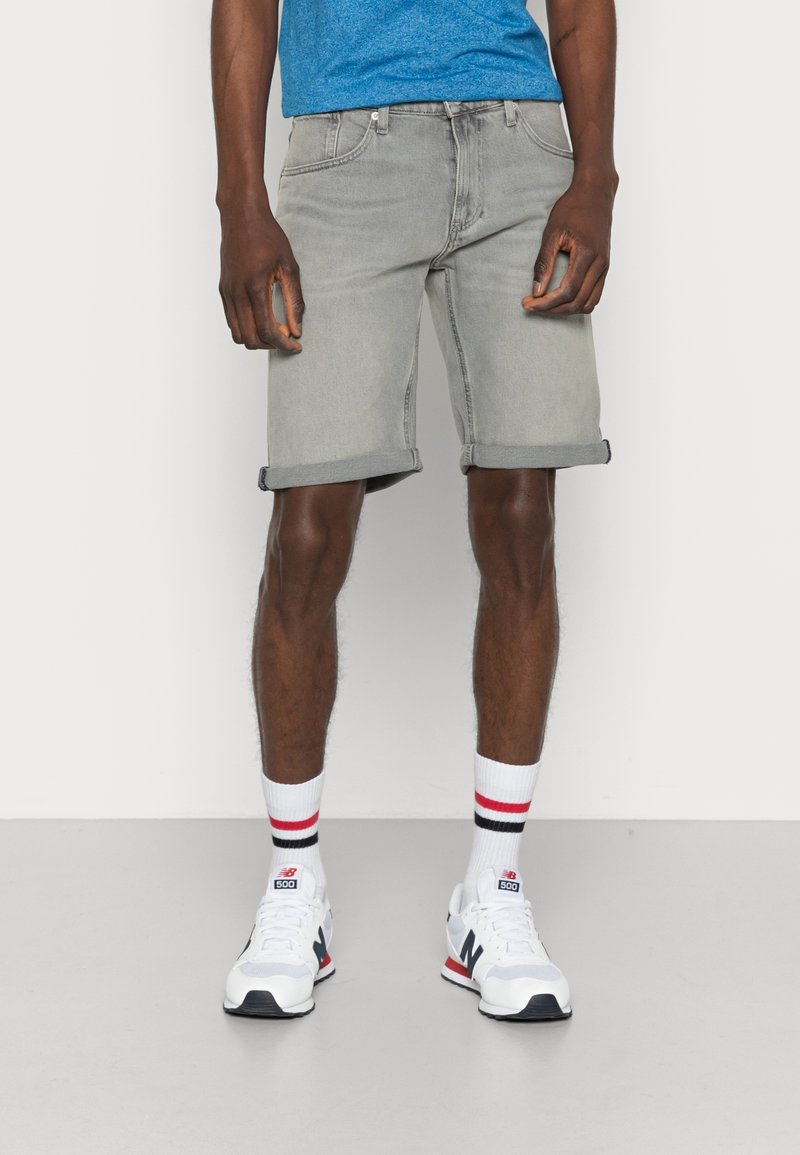 Pantalones cortos de mezclilla gris con puños enrollados, combinados con calcetas blancas con rayas rojas y negras, y zapatillas blancas. Camiseta azul visible.