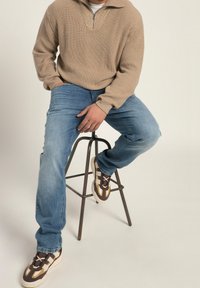 Maglione beige con zip a 1/4, jeans in denim blu e scarpe da ginnastica marroni con dettagli beige. Modello seduto su uno sgabello di metallo, abbigliamento casual.