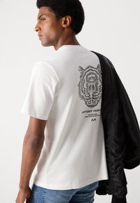 T-shirt bianca in cotone con grafica di un leone nera sulla schiena, accompagnata dal testo "ANTONY MORATO". Modello abbinato a una giacca bomber nera.