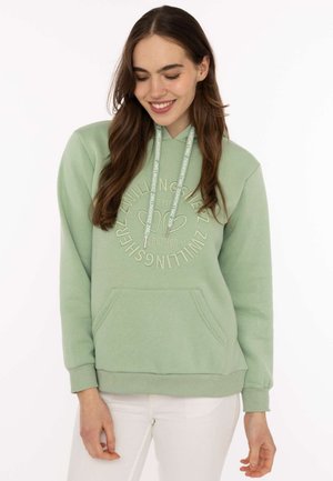 Junge Frau mit langen braunen Haaren, die einen hellgrünen Hoodie mit Text und weißen Hosen trägt, lächelnd und nach unten schauend.