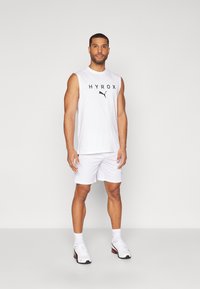 Biela bezrukávová športová tangá s logom "HYROX", skombinovaná s bielymi šortkami, ukazujúca hladkú text�úru látky a uvoľnený strih.