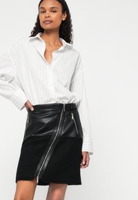 Μαύρη φούστα από δέρμα και υφασμάτινη υφή, με διαγώνιο φερμουάρ μπροστά, συνδυασμένη με λευκή πουκαμίσα με ρίγες.