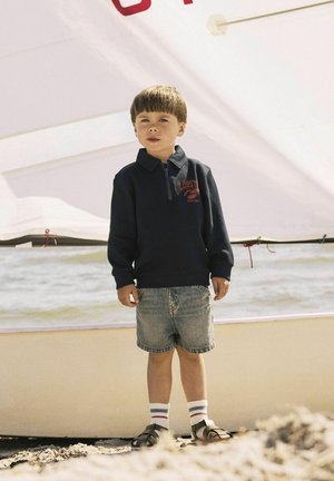 Dunkelblaue Zip-Up-Sweatjacke mit rotem "LOBSTER"-Logo, kombiniert mit blauen Jeansshorts und gestreiften Socken, getragen mit braunen Sandalen am Strand.