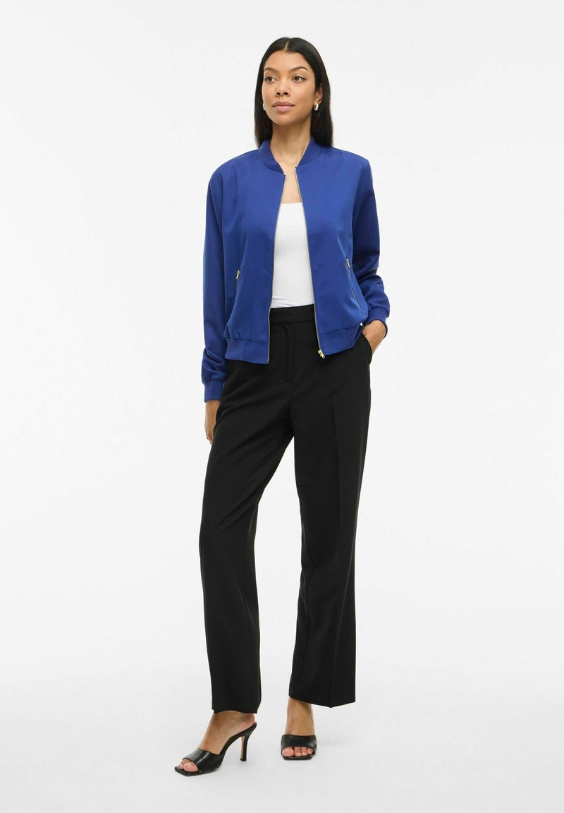Femme portant une veste zippée bleu royal, un haut blanc, un pantalon noir à jambes droites et des sandales à talons ouverts noires, se tenant avec une main dans sa poche.