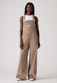 Brown corduroy overalls with wide legs, featuring adjustable shoulder straps and front pocket. Worn over a white crew-neck top.