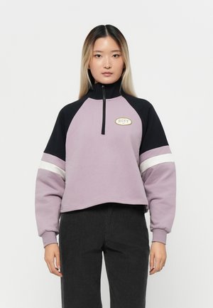 Lilla avkortet hettegenser med høy krage, svarte ermer og hvite aksentstriper. Har et gull "Roxy"-logo på brystet.