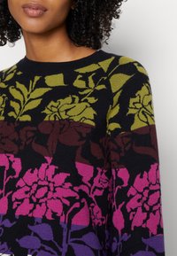 Femme portant un pull multicolore avec des motifs floraux verts, bruns, roses et violets sur un fond noir.