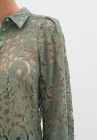 Krajková blúzka v tlmenej zelenej farbe s kvetinovými a paisley vzormi, s gombíkovým golierom a polopriesvitnou textúrou. Dlhé rukávy.