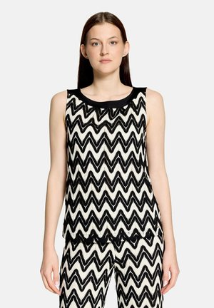 Femme portant un haut sans manches et un pantalon avec un motif zigzag noir et blanc sur fond gris clair.