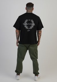 Svart bomullströja med vitt grafiskt logo och lagerkrans på ryggen, kombinerad med gröna cargobyxor och vita sneakers.