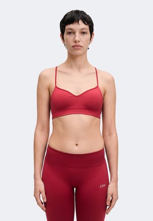 Jeune femme aux cheveux courts et foncés, portant un brassard de sport rouge et un legging taille haute, debout sur un fond clair uni.