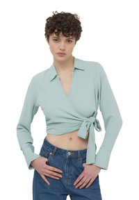 Blusa avvolgente verde menta con maniche lunghe e dettaglio a fiocco in vita. Presenta un colletto e polsini ripiegati. Abbinata a jeans a vita alta.