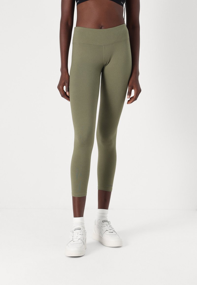 Oliven grønne leggings med høy midje, kort design og glatt tekstur. Kombinert med hvite joggesko og synlige hvite sokker.