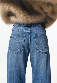 Vue arrière d'un jean en denim bleu avec des clous métalliques en forme d'étoile sur les poches, porté avec un manteau en fourrure marron clair.