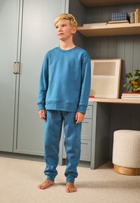 Plavi komplet od pamučne trenirke s rebrastim manšetama, opuštenim krojem i mekanom teksturom. U pozadini sobe nalaze se police i biljka.