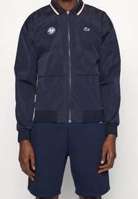 Man som bär en marinblå zip-up jacka med Roland Garros- och Lacoste-logotyper, tillsammans med matchande marinblå shorts, stående mot vit bakgrund.