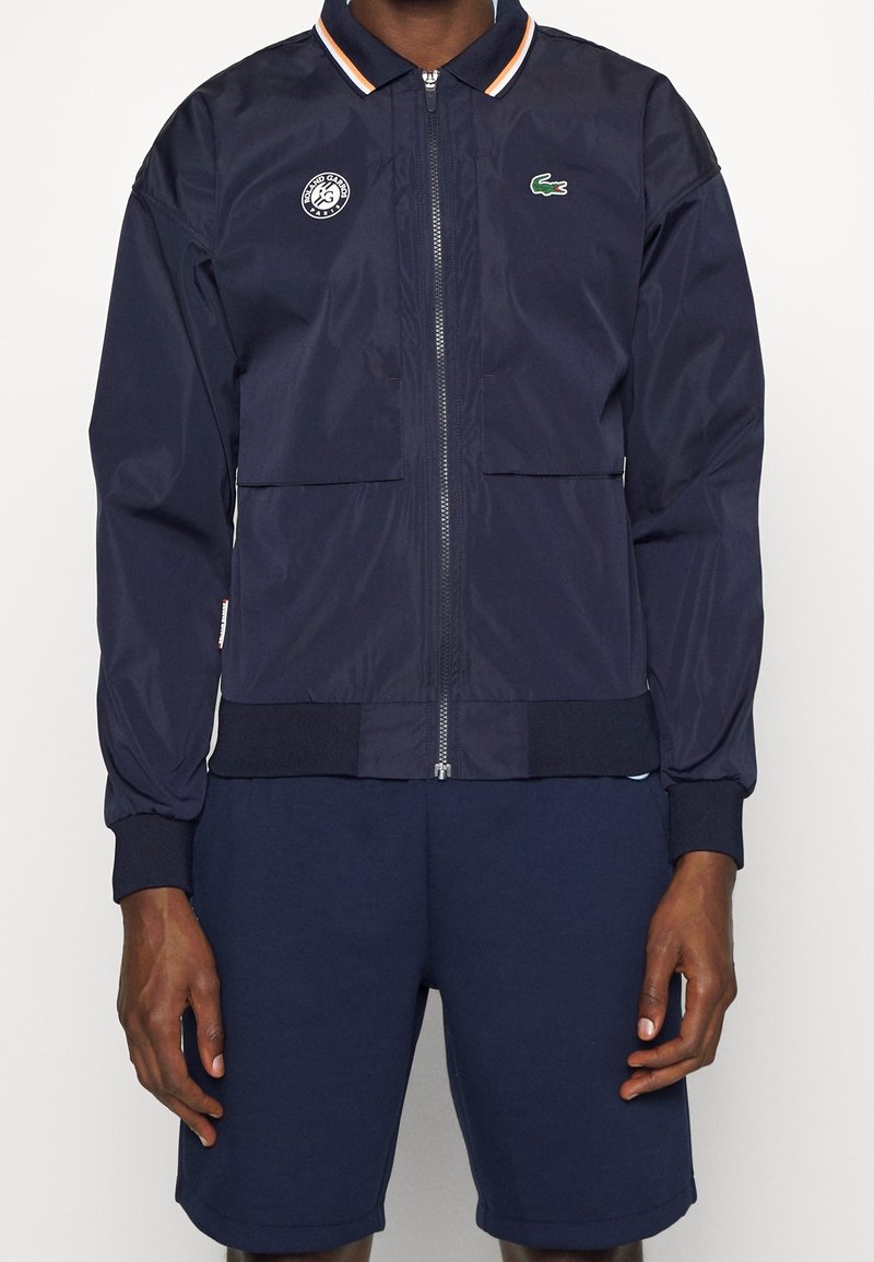 Man som bär en marinblå zip-up jacka med Roland Garros- och Lacoste-logotyper, tillsammans med matchande marinblå shorts, stående mot vit bakgrund.