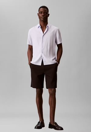 Uomo in piedi con le mani in tasca che indossa una camicia bianca a maniche corte con bottoni, pantaloncini neri e mocassini neri su uno sfondo grigio.
