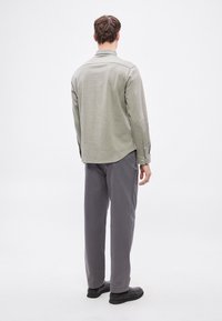 Svetlo zelena srajca z ovratnikom, ravnim spodnjim robom in gumbi na manšetah, kombinirana s sivimi hlačami in črnimi čevlji, videna od zadaj.