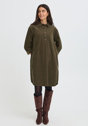 Femme aux longs cheveux foncés portant une robe vert olive boutonnée, des collants noirs et des bottes marron montant jusqu'aux genoux, debout les mains dans les poches.