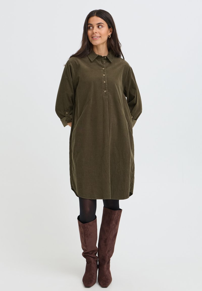 Femme aux longs cheveux foncés portant une robe vert olive boutonnée, des collants noirs et des bottes marron montant jusqu'aux genoux, debout les mains dans les poches.