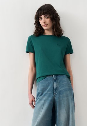 Tânără cu păr creț închis la culoare până la umeri poartă un tricou verde cu mâneci scurte și blugi albaștri largi pe un fundal simplu.