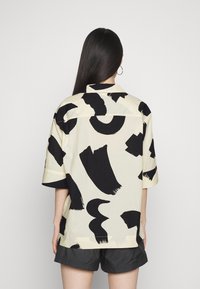 Camicia a maniche corte color crema chiaro con audaci motivi a pennellate nere. Presenta un colletto e una vestibilità rilassata.