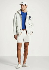 Νεαρός άνδρας που φοράει λευκό καπέλο, λευκό μπουφάν, μπλε πουκάμισο, λευκά σορτς με καφέ ζώνη και λευκά αθλητικά παπούτσια, στέκεται απέναντι από ένα απλό φόντο.