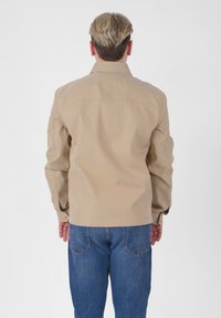 Giacca beige con colletto classico, maniche lunghe e polsini con bottoni; abbinata a jeans blu. Tessuto liscio, design sobrio, vestibilità slim.