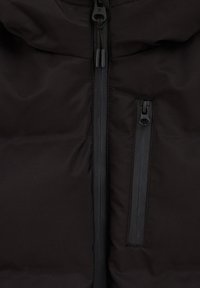 Juodas izoliacinis rūbas su centriniu užtrauktuku, dviem užtrauktukais užsegamomis kišenėmis ir lygiu, matinio paviršiaus apdaila. Sukurtas pūstai siluetui.