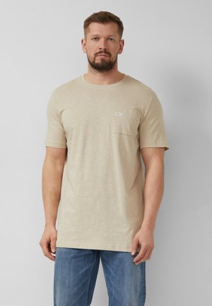 Mężczyzna z brodą w beżowym krótkim rękawem koszulką z kieszonką na piersi i niebieskich dżinsach, stojący na jednolitym szarym tle.