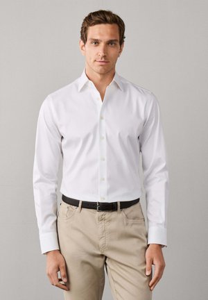 Man draagt een wit overhemd met lange mouwen en knopen, gestopt in beige broek met een zwarte riem, staand voor een effen achtergrond.