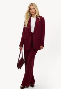 Bordeaux tailor-made pak met een lange blazer en wijde broekspijpen. Gecombineerd met een witte top en een gestructureerde, bijpassende handtas.