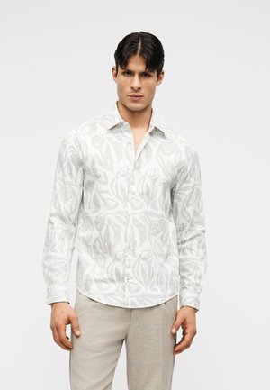 Meessõltuuval modellil on seljas valge nööbitav särk helehallide lillemustriga ja beežid püksid, ta seisab ühevärvilise valge tausta ees.