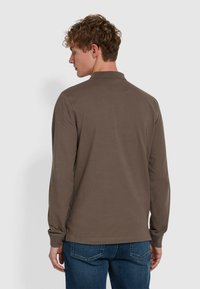 Langärmliges, braunes Poloshirt aus weichem Stoff mit Standardkragen, schmaler Passform und sichtbaren Nähdetails. Kombiniert mit blauen Jeans.