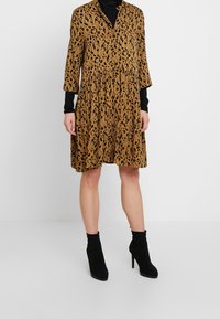 Braun gepunktetes Kleid mit V-Ausschnitt, Dreiviertelärmeln und einem ausgestellten Rock. Kombiniert mit schwarzen Stiefeletten und einem schwarzen Rollkragenpullover.
