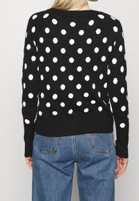 Schwarzer Pullover mit weißen Polka-Dots, gerippte Bündchen und Saum, mittelgrobe Strickstruktur, kombiniert mit hellblauen, hochgeschnittenen Jeans.