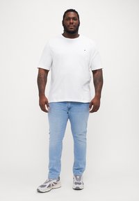 Hvit bomulls t-skjorte, avslappet passform, kombinert med lyseblå denim jeans og grå-detaljerte joggesko, stående mot en ensfarget bakgrunn.