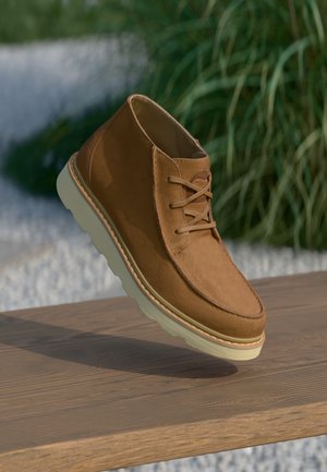 Stivaletto alla caviglia in camoscio marrone con suola e lacci beige chiaro, mostrato fluttuante sopra una superficie in legno con piante verdi sullo sfondo.