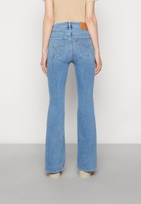 Jean évasé bleu en denim, avec une taille haute, des coutures visibles et deux poches arrière, avec une étiquette logo en cuir fauve sur la ceinture.