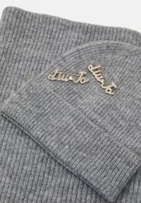 Beanie grigio in maglia con texture a costine, dotata di una spilla in tono oro decorata con cristalli trasparenti, che forma la scritta "liuta" in carattere corsivo.