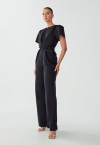 Zwarte jumpsuit met korte, geplooide mouwen, een wikkellijn en een gestrikte taille. Heeft rechte, brede pijpen en een soepele stoftextuur.