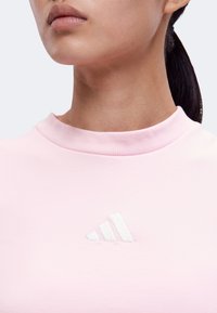 Νεαρή γυναίκα με ανοιχτό ροζ τοπ Adidas με λευκό λογότυπο, τα μαλλιά δεμένα πίσω, που δείχνει τον λαιμό και το κάτω μέρος του προσώπου σε απλό φόντο.