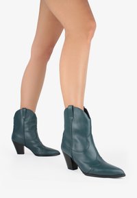 Botas de cowboy de cuero verde hasta el tobillo con punta afilada, tacón inclinado y costuras decorativas. Textura suave y lengüetas para facilitar su uso.
