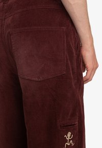 Burgundy corduroy pants with vertical ribbing; features back pocket and side pocket with a spiral figure embroidery detail.
