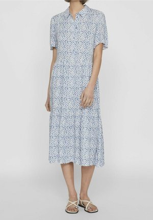 Kvinde iført en lyseblå blomsterprint midi-kjole med korte ærmer og krave, kombineret med hvide flade sandaler med stropper.